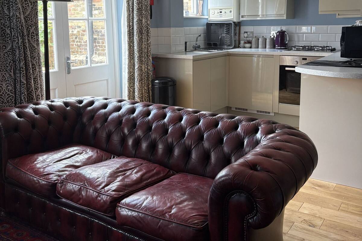 A living room with a dark brown leather sofa and a patterned rug. A kitchen with white cabinets and a gas stove is visible in the background.