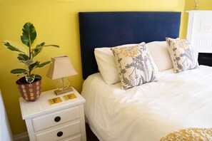 A bed with white bedding and patterned pillows, a bedside table with a lamp and a potted plant, and a yellow wall.