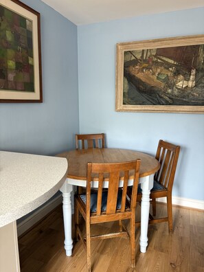 A dining area with a round wooden table and four chairs, a painting on the wall, and a framed picture above the table.