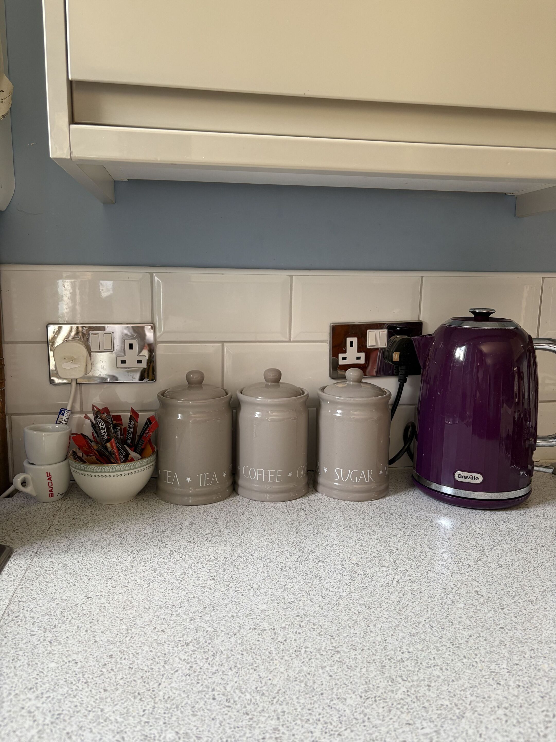 A kitchen counter with storage jars labeled tea, coffee, and sugar, a coffee maker, and a mug.