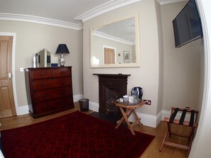 Ein Zimmer mit Kamin, einem hölzernen Schminktisch, einem roten Teppich mit Muster sowie einem Spiegel und einem an der Wand befestigten Fernseher.