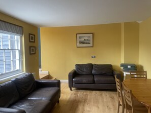Un salon avec un canapé, une table à manger avec des chaises, une fenêtre avec des stores et un tableau encadré au mur.