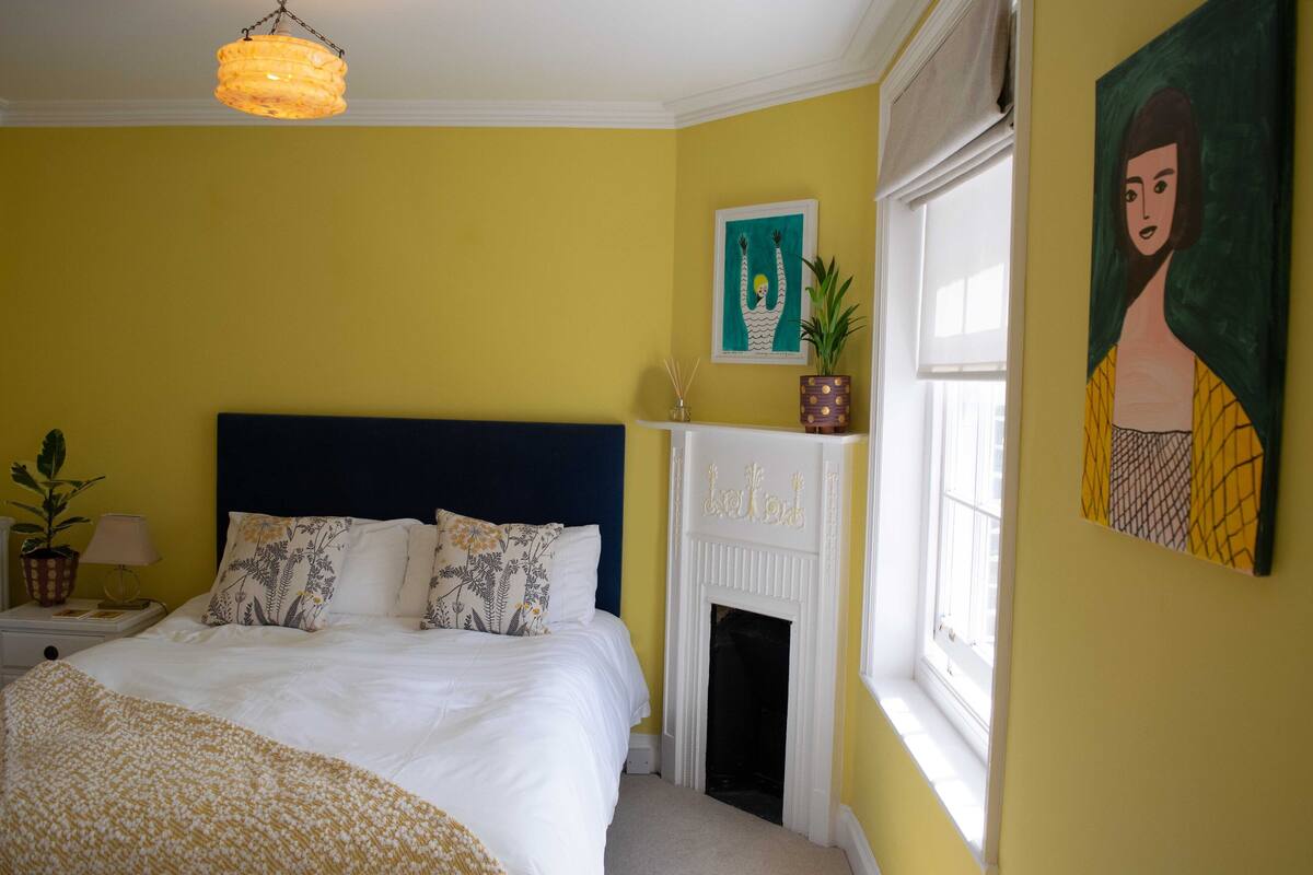 A bedroom with a bed, a nightstand, a lamp, a fireplace, and artwork on the walls.