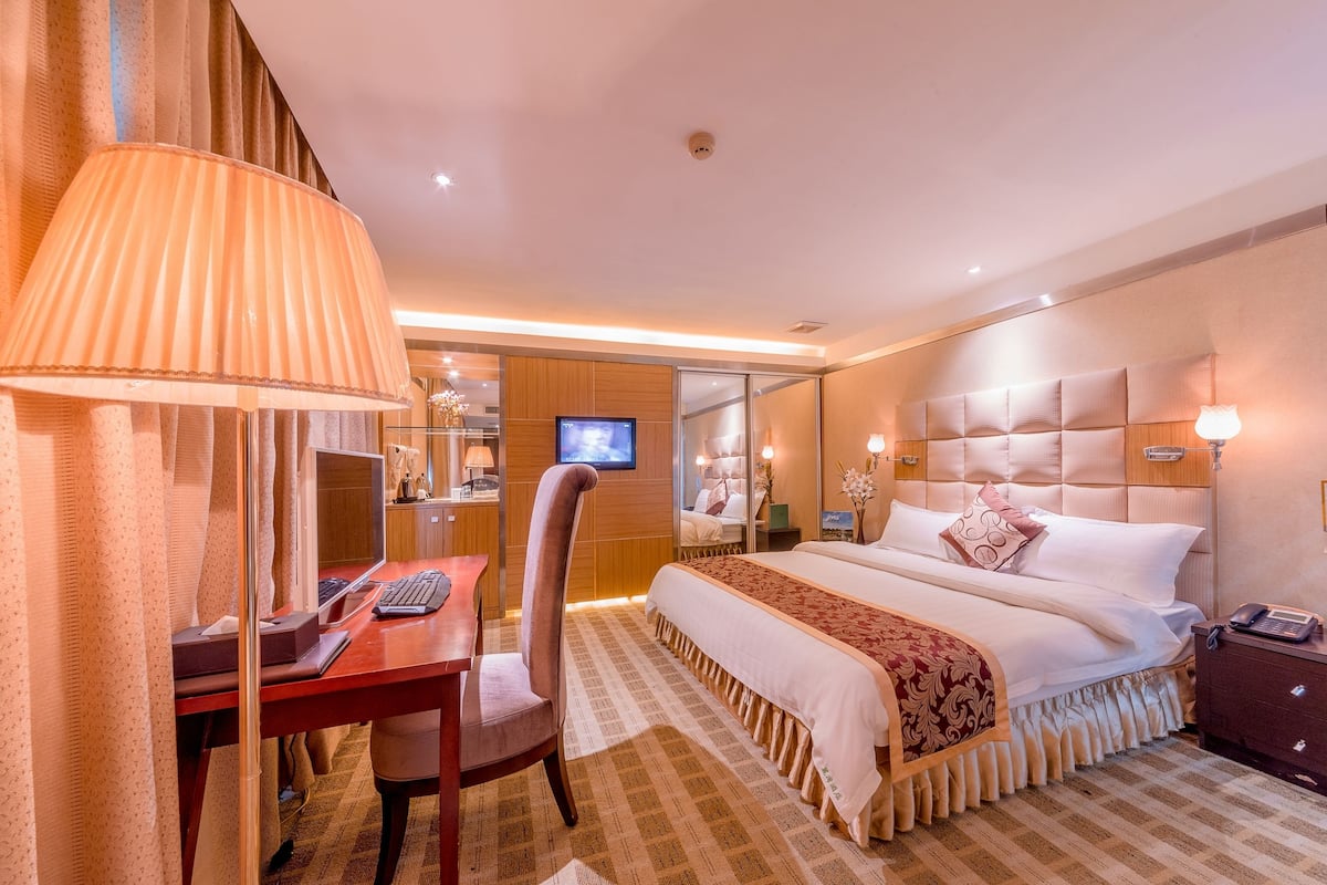 A hotel room with a bed, a desk with a computer, a chair, a TV, and a bedside table with a lamp.