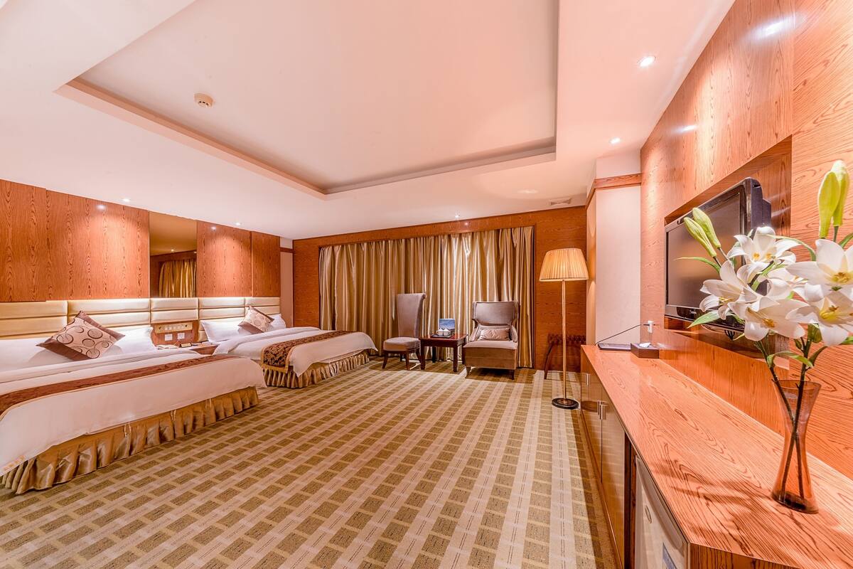 A hotel room with two beds, a wooden reception desk, a desk lamp, a chair, and a vase with flowers.