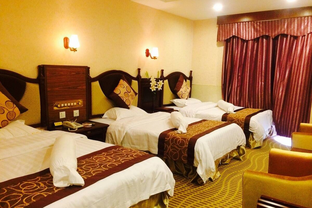 A hotel room with three beds, each with a wooden headboard and white linens. There are two bedside tables with lamps and a telephone. The room has a patterned carpet and a window with red curtains.