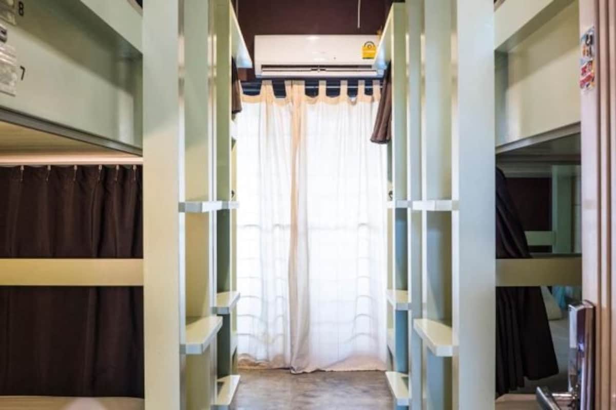 Un couloir étroit, avec des lits superposés de chaque côté, un rideau au fond et un appareil de climatisation au plafond.