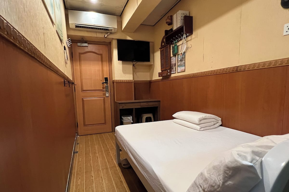 A hotel room with a wooden door, a bed with white linens, a small table, a wall-mounted TV, and an air conditioning unit.
