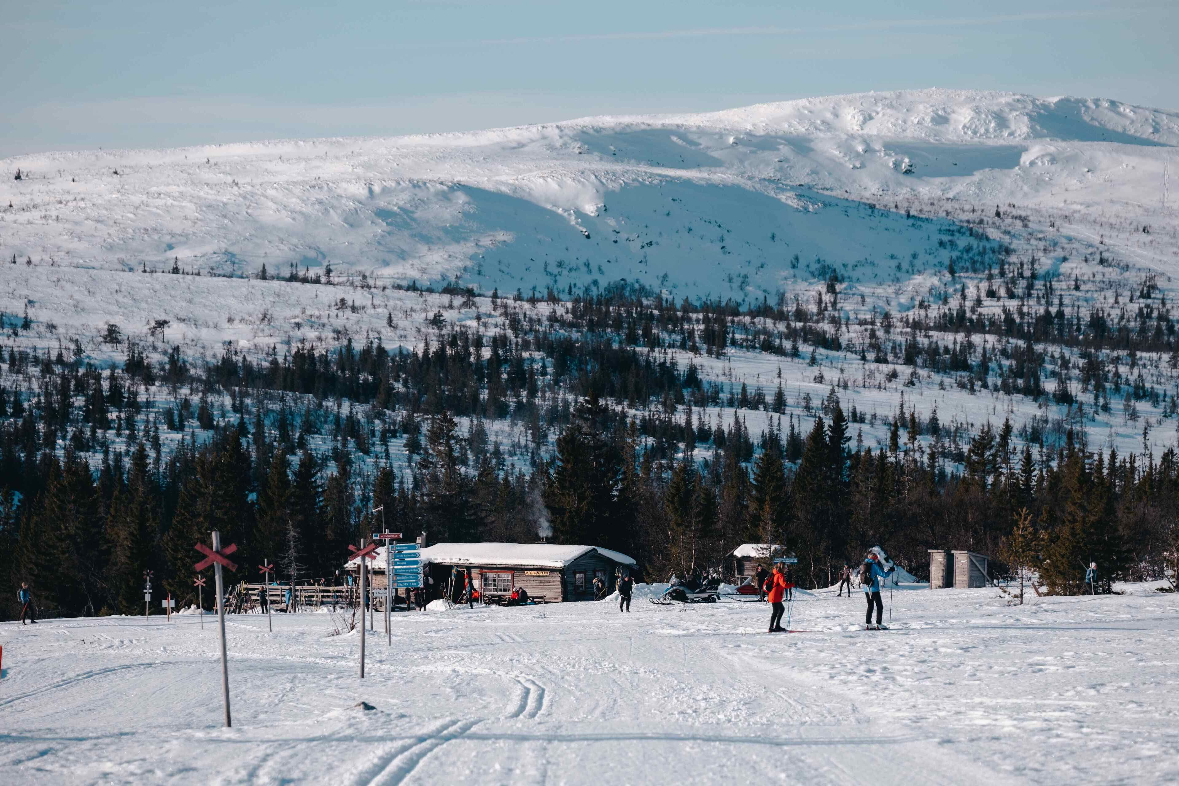 Et snøklædt skiområde med en hytte, skiløjper og et bjerg i baggrunden.