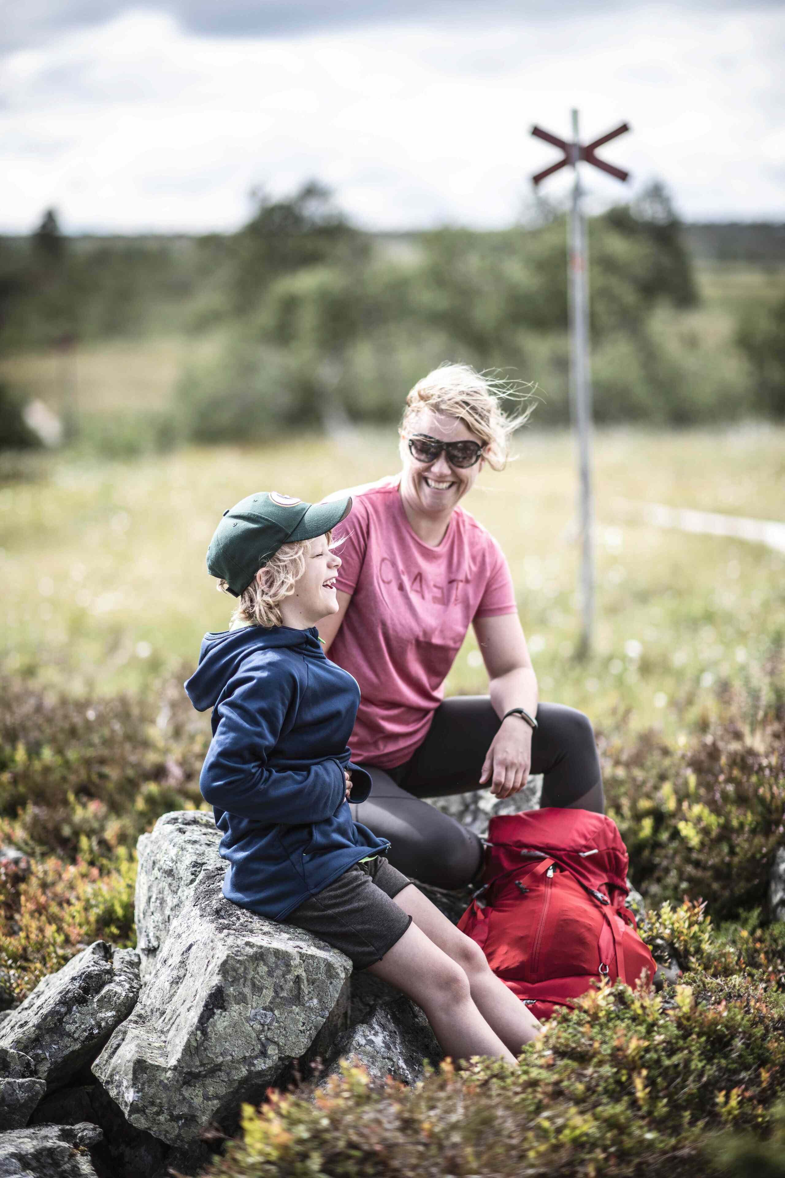 Twee personen zitten op een rots, met een rode rugzak en een overwegborduurwerk op de achtergrond.