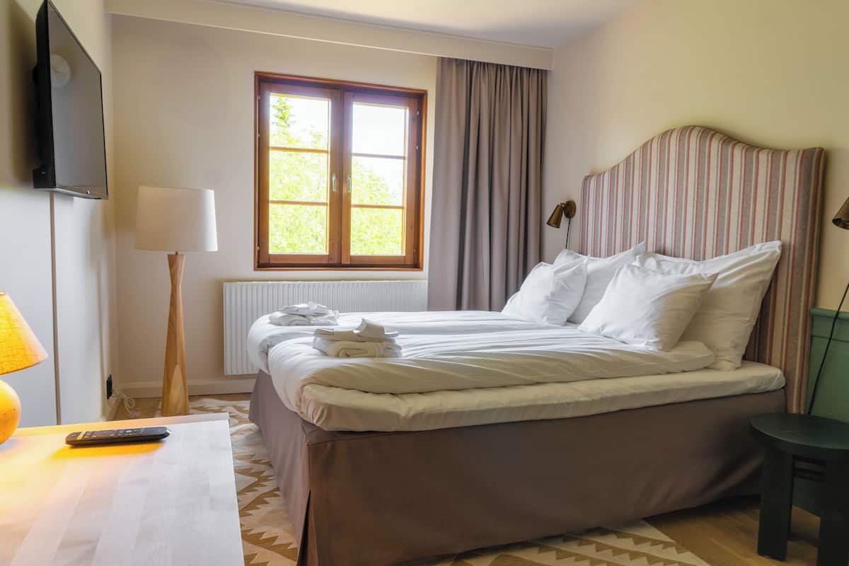 Een hotelkamer met een bed, een bureau met lamp, een televisie, een raam met gordijnen en een radiator.