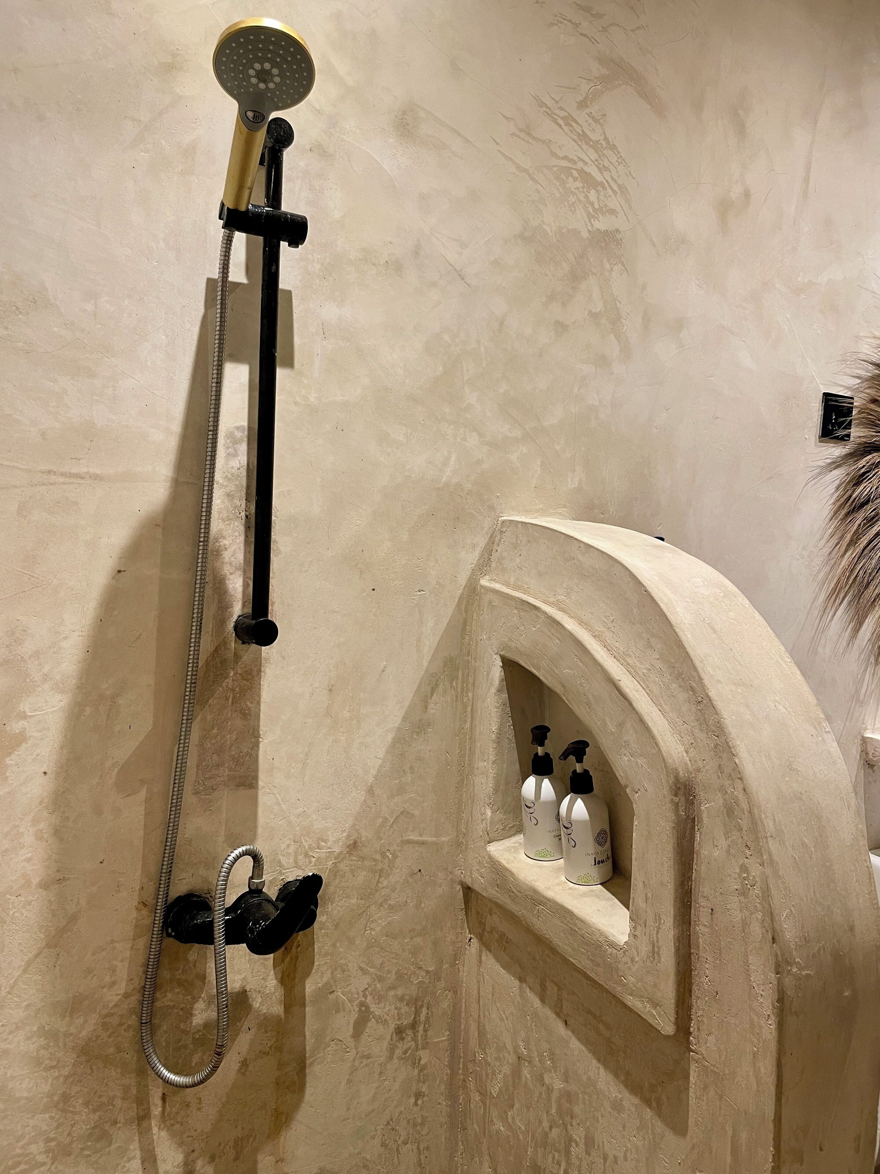 A shower with a handheld showerhead and a wall-mounted faucet. There is a small shelf with two bottles of shampoo and conditioner.