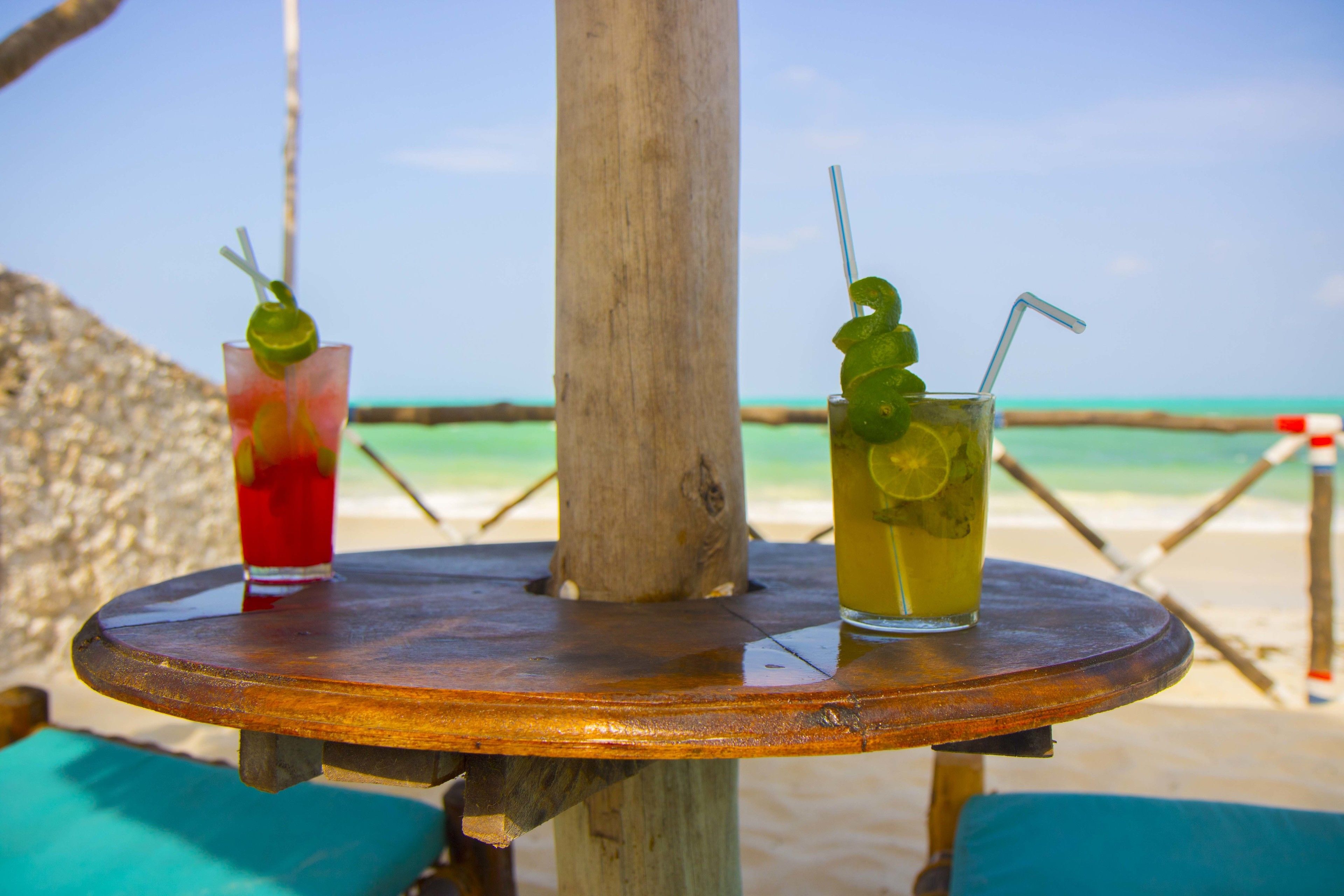 Dos bebidas sobre una mesa de madera, con vista a la playa y al océano de fondo.
