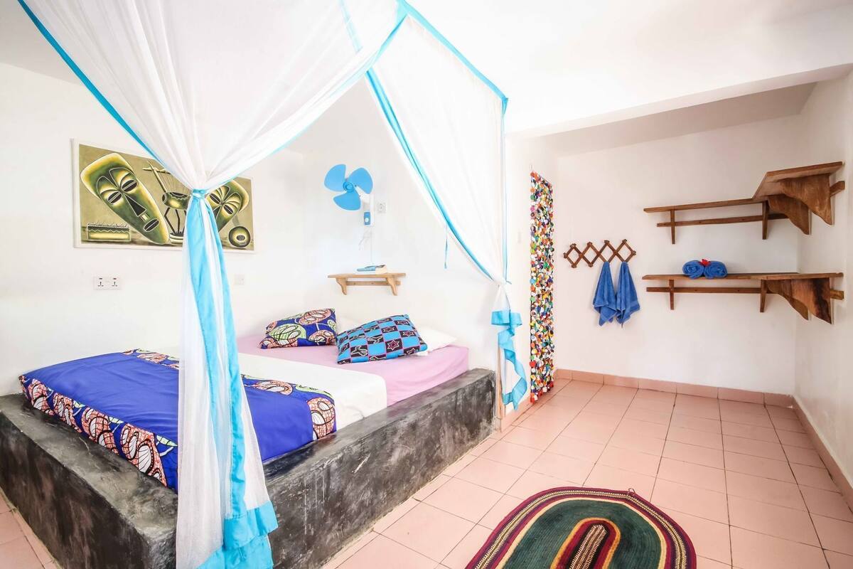 A room with a bed under a canopy, a wall-mounted fan, a shelf with towels, and a decorative wall piece.