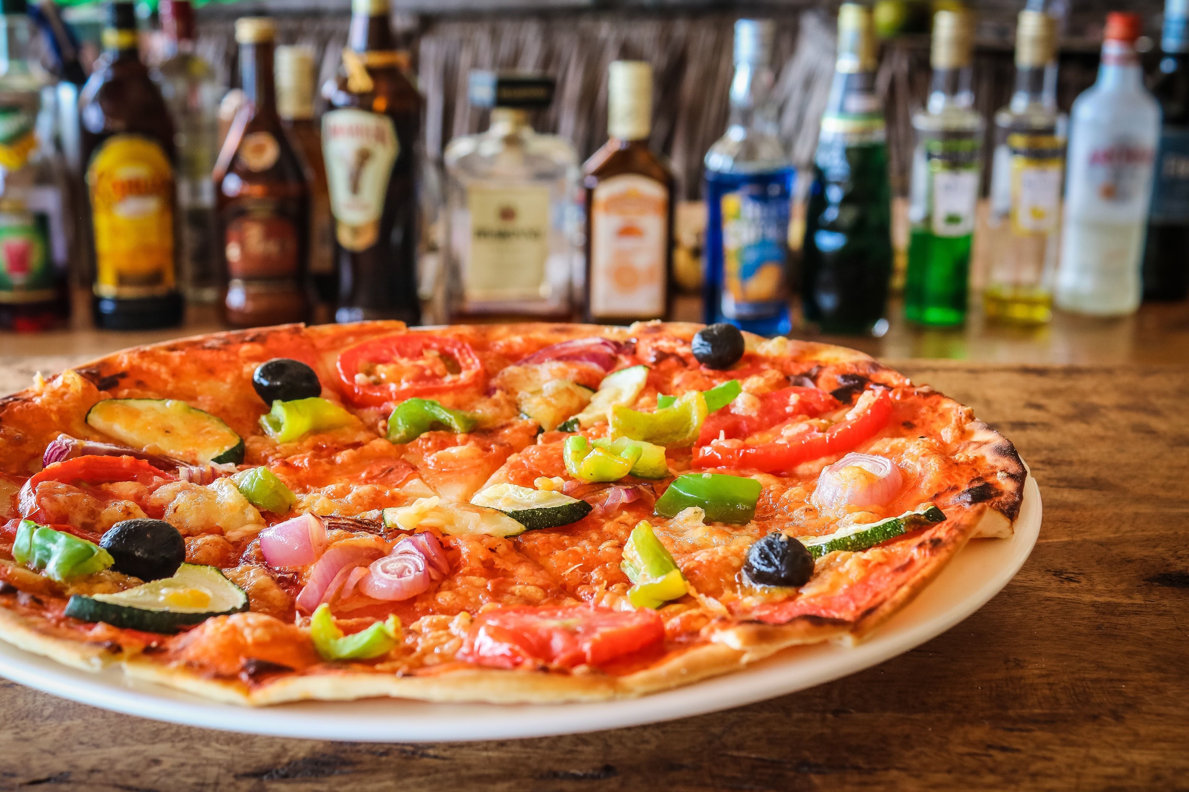 Eine Pizza mit verschiedenen Belägen auf einem weißen Teller, platziert auf einem Holztisch. Im unscharfen Hintergrund sind verschiedene Flaschen zu sehen.