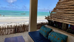 Vista a la playa desde un espacio cubierto con valla de madera, estructura de madera y cama con almohadones estampados.