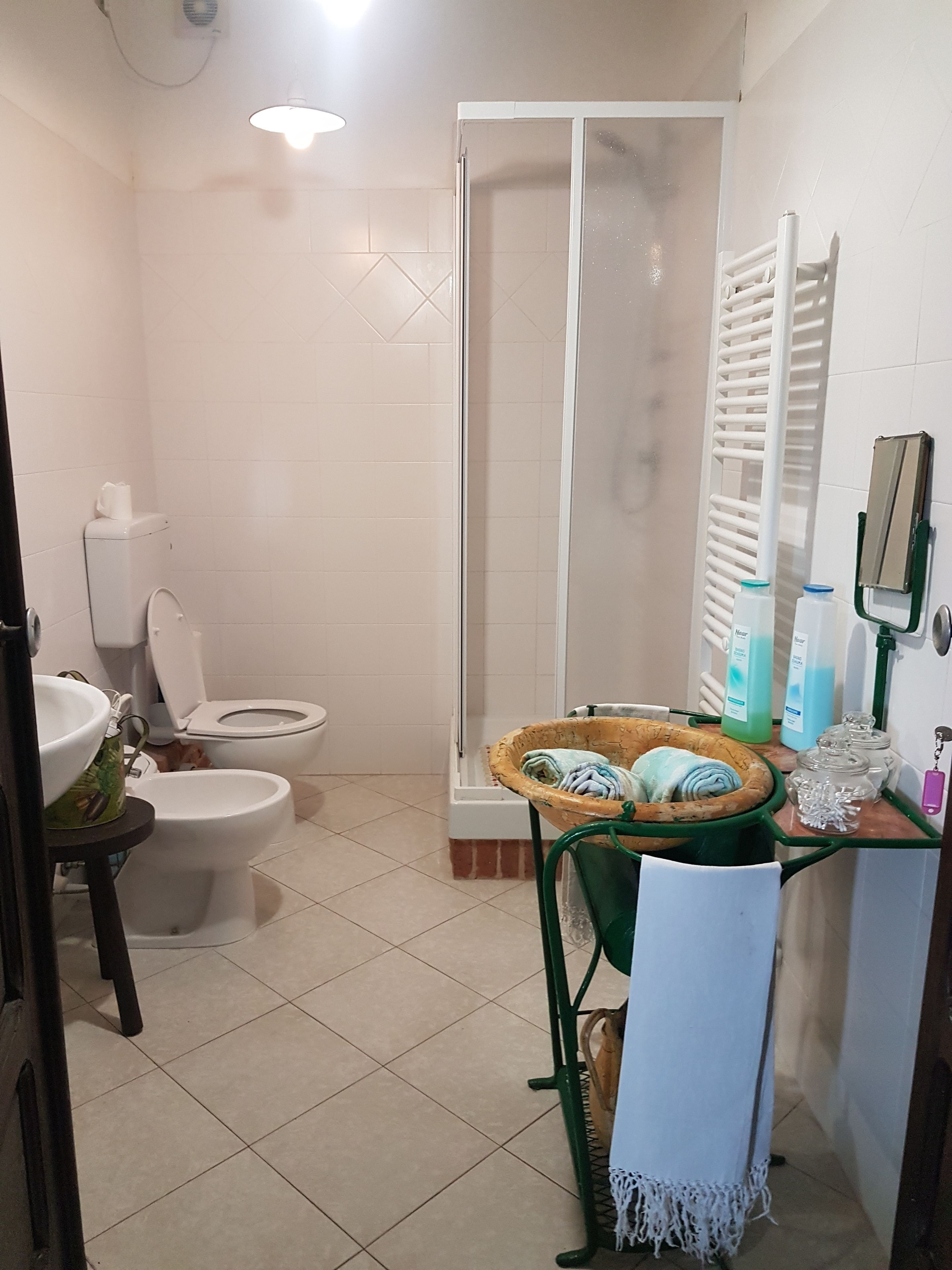 A bathroom with a toilet, bidet, and shower. There is a sink with a bowl, a green towel rack, and a basket with towels.