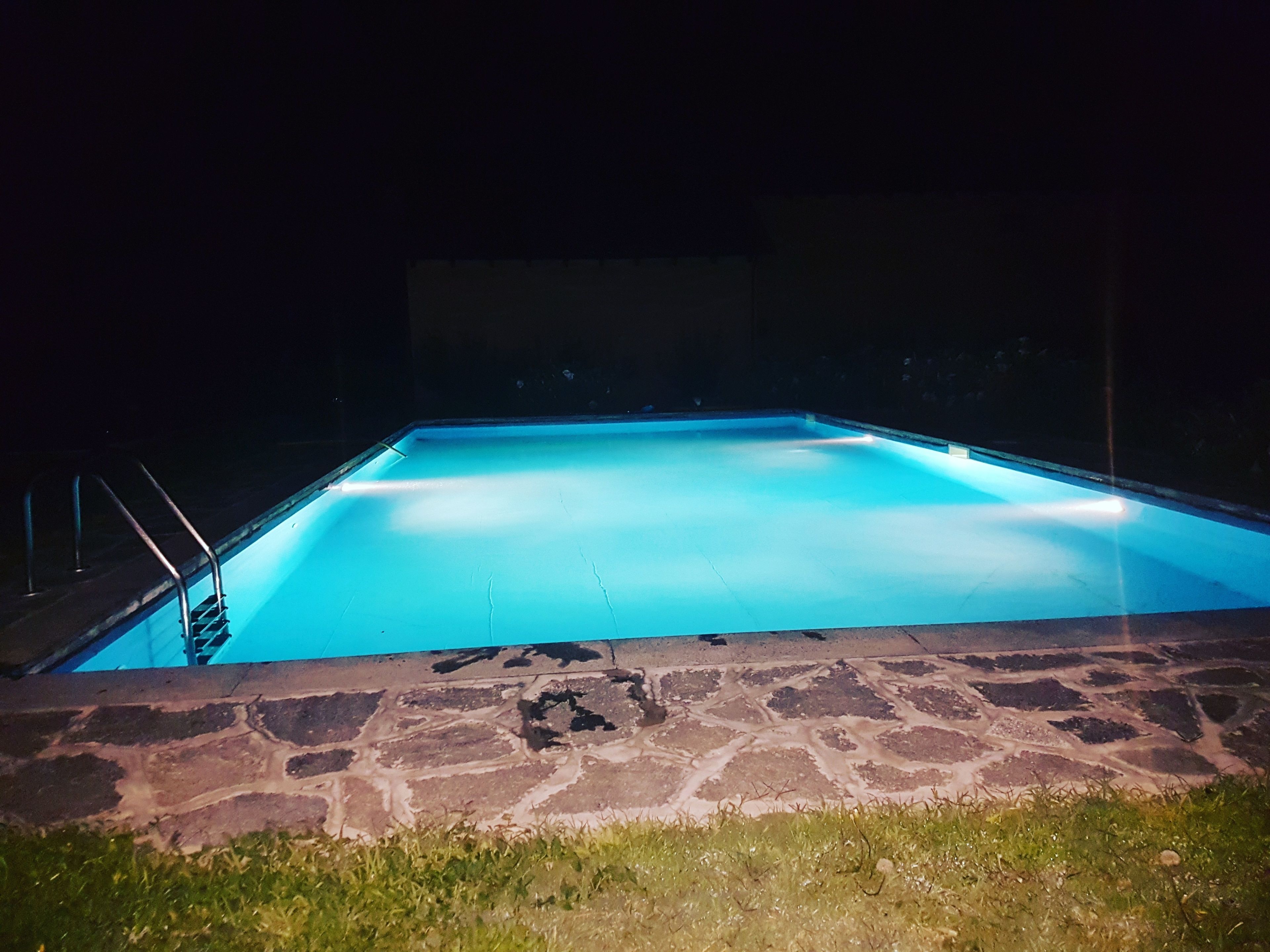 A rectangular swimming pool with a stone border and metal handrails, illuminated by blue lighting.