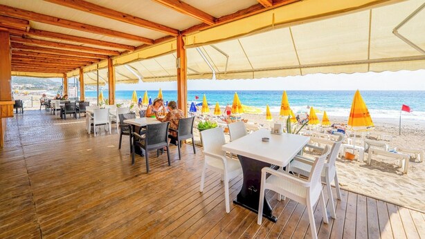 Un restaurante al aire libre con suelo de madera y mesas, con vistas a una playa con sombrillas y gente.