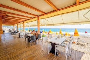 Un restaurante al aire libre con suelo de madera y mesas, con vistas a una playa con sombrillas y gente.