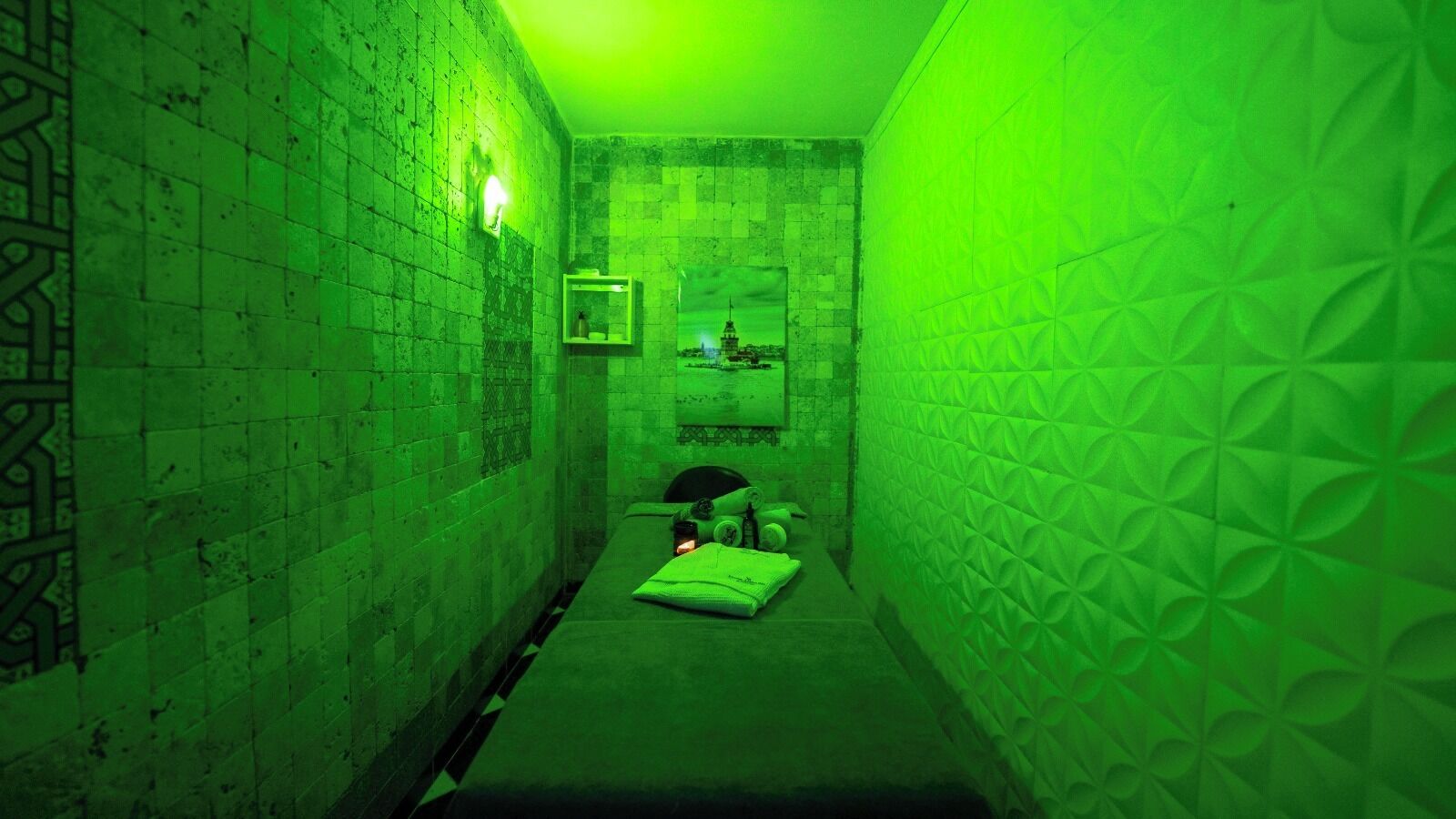 A narrow hallway with a green hue, patterned walls, a bed with pillows, and a small table with items.