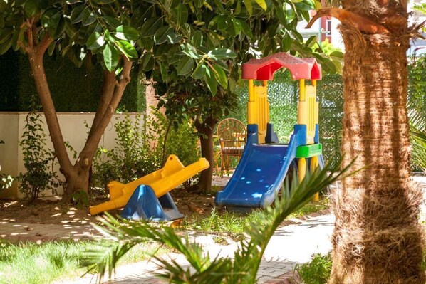 A playground with a slide and swings, surrounded by trees and a brick pathway. - Galaxy Beach Hotel (Alanya)