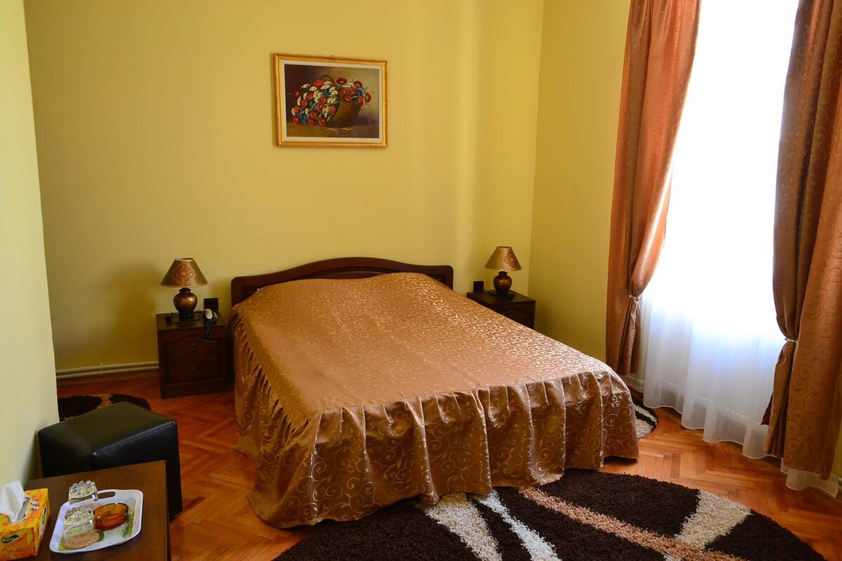 A neatly arranged bedroom with a bed, two bedside tables with lamps, a small table with a plate of food, and a painting on the wall.