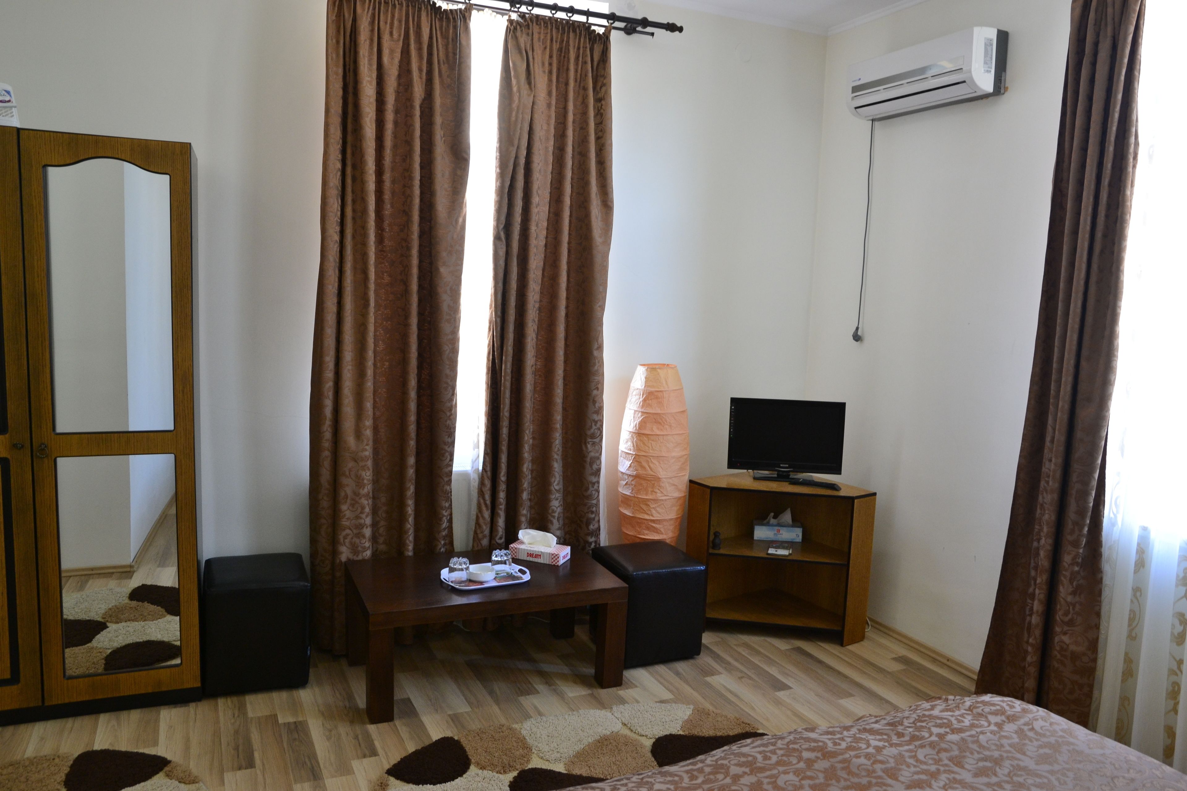 Une chambre d’hôtel avec une armoire en bois, une petite table avec un téléphone et une lampe, un téléviseur posé sur un meuble en bois, et un climatiseur fixé au mur.