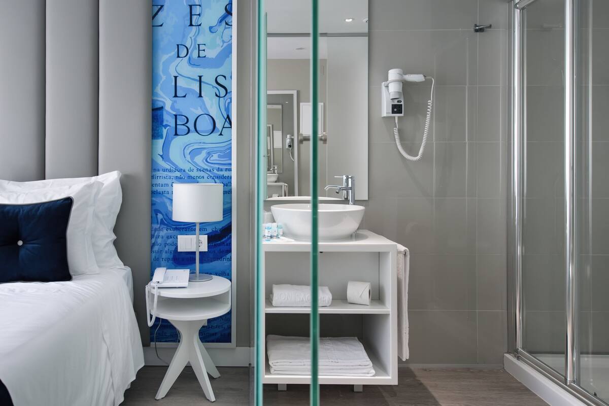 Ein modernes Hotelzimmer mit Bett, Nachttisch mit Lampe, Badezimmer mit Waschbecken und Dusche.