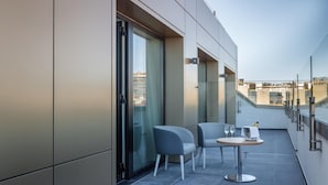 Un balcón moderno con dos sillas y una mesita pequeña, grandes puertas correderas de vidrio y vistas a la ciudad.