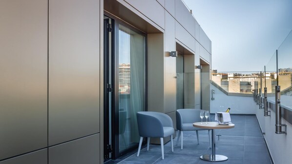 A modern balcony with two chairs and a small table, large sliding glass doors, and a city view.