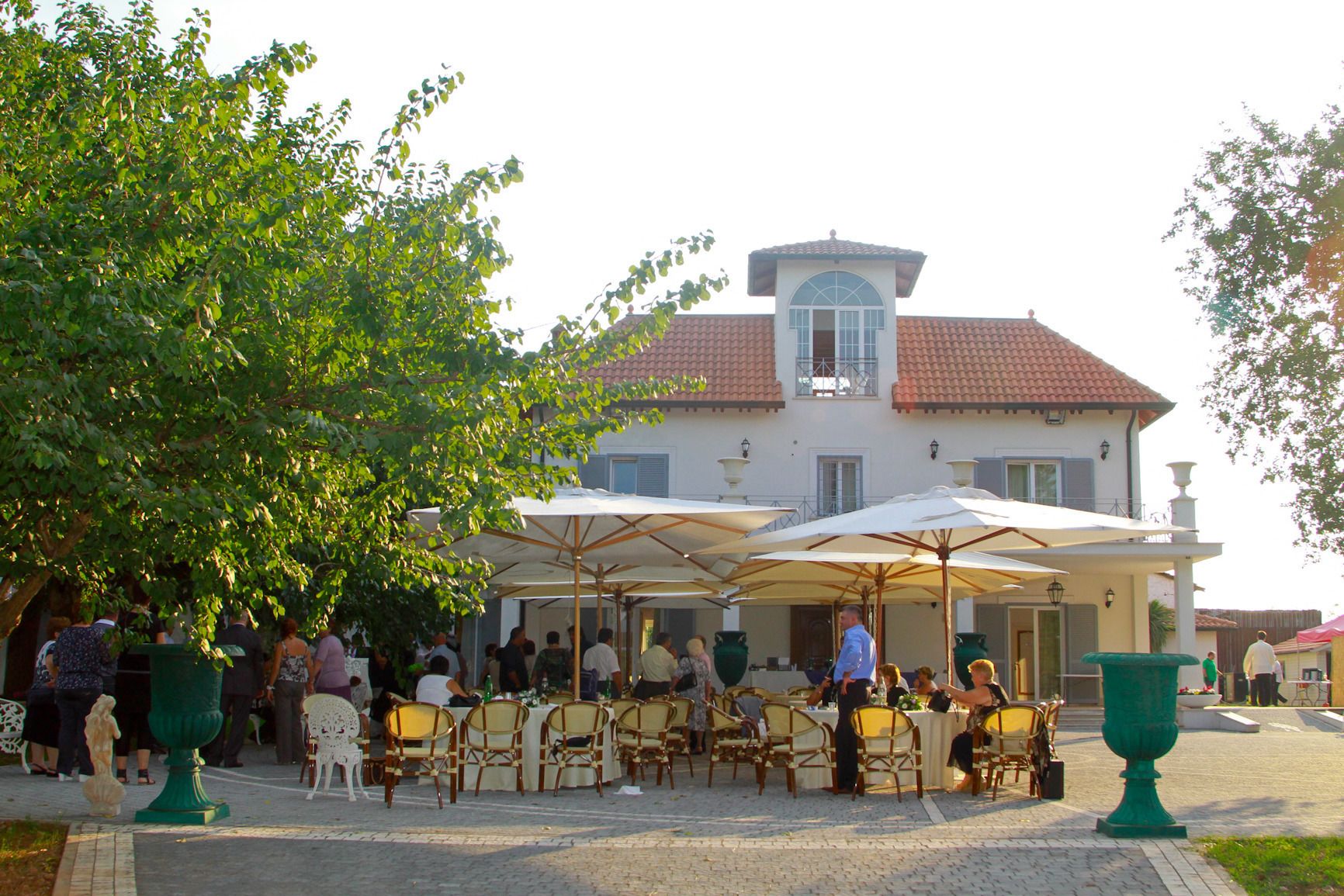 An outdoor dining area with tables and chairs, surrounded by trees and a building with a tiled roof.
