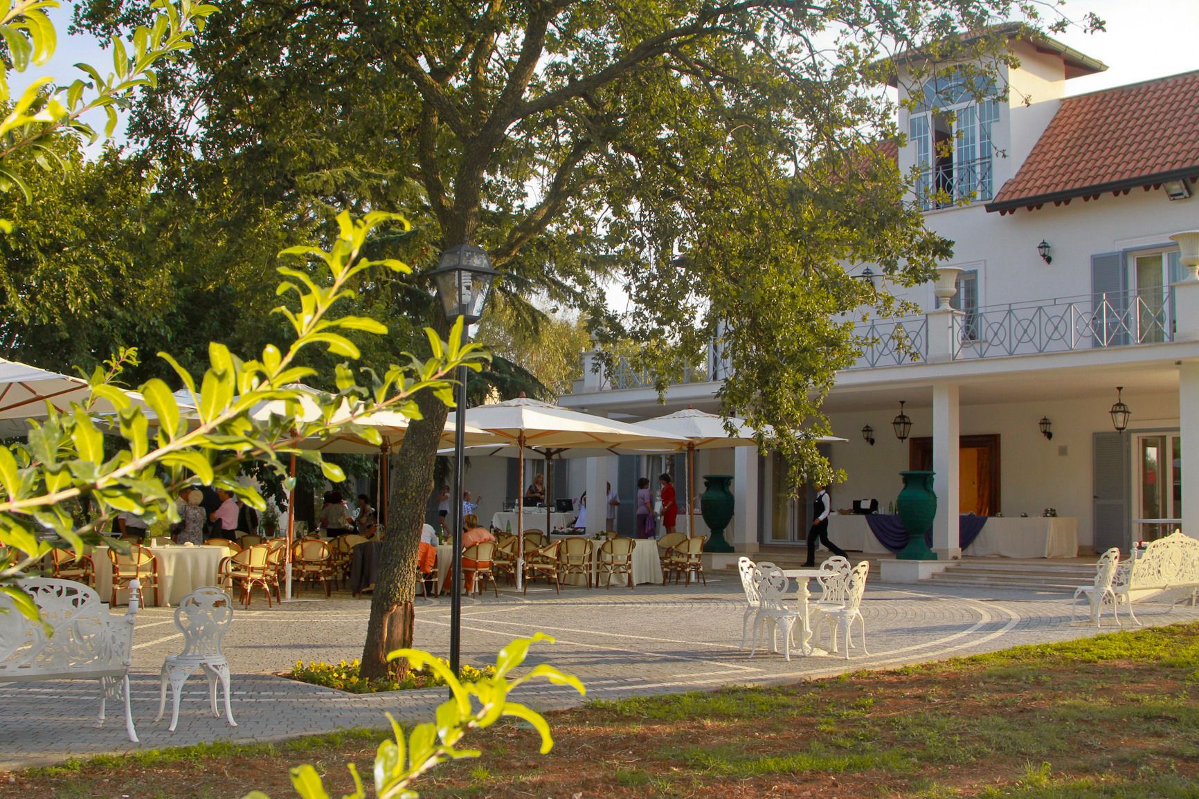 An outdoor café area with tables and chairs, surrounded by trees and a building with a balcony.