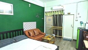 A hotel room with a bed, a brown leather chair, a small wooden table, a green wall, and a door with a curtain. - The Backpacker Station (Pak Kret)