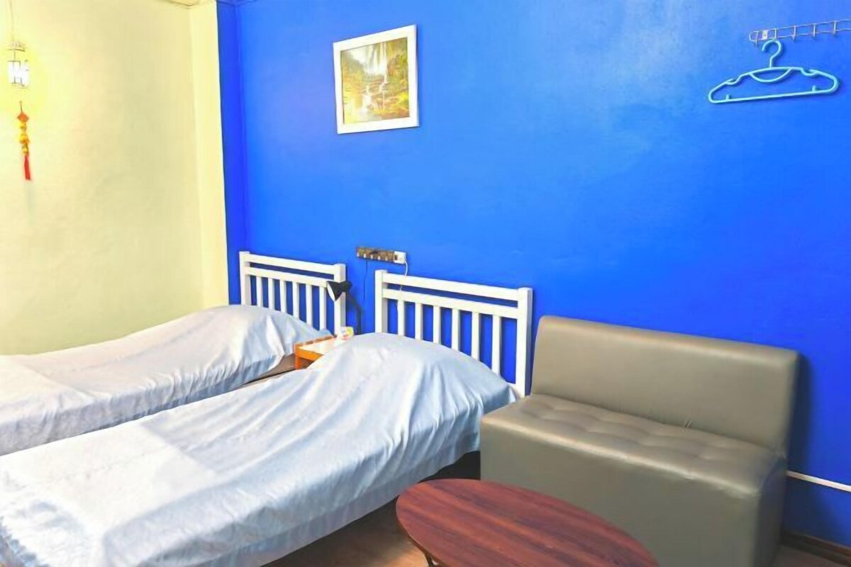 A room with two single beds, a small table, a chair, and a blue wall with a picture and a hanger.
