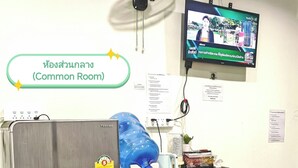 A common room with a refrigerator, a large blue water dispenser, a table with various items, a standing fan, and a television mounted on the wall. - The Backpacker Station (Pak Kret)