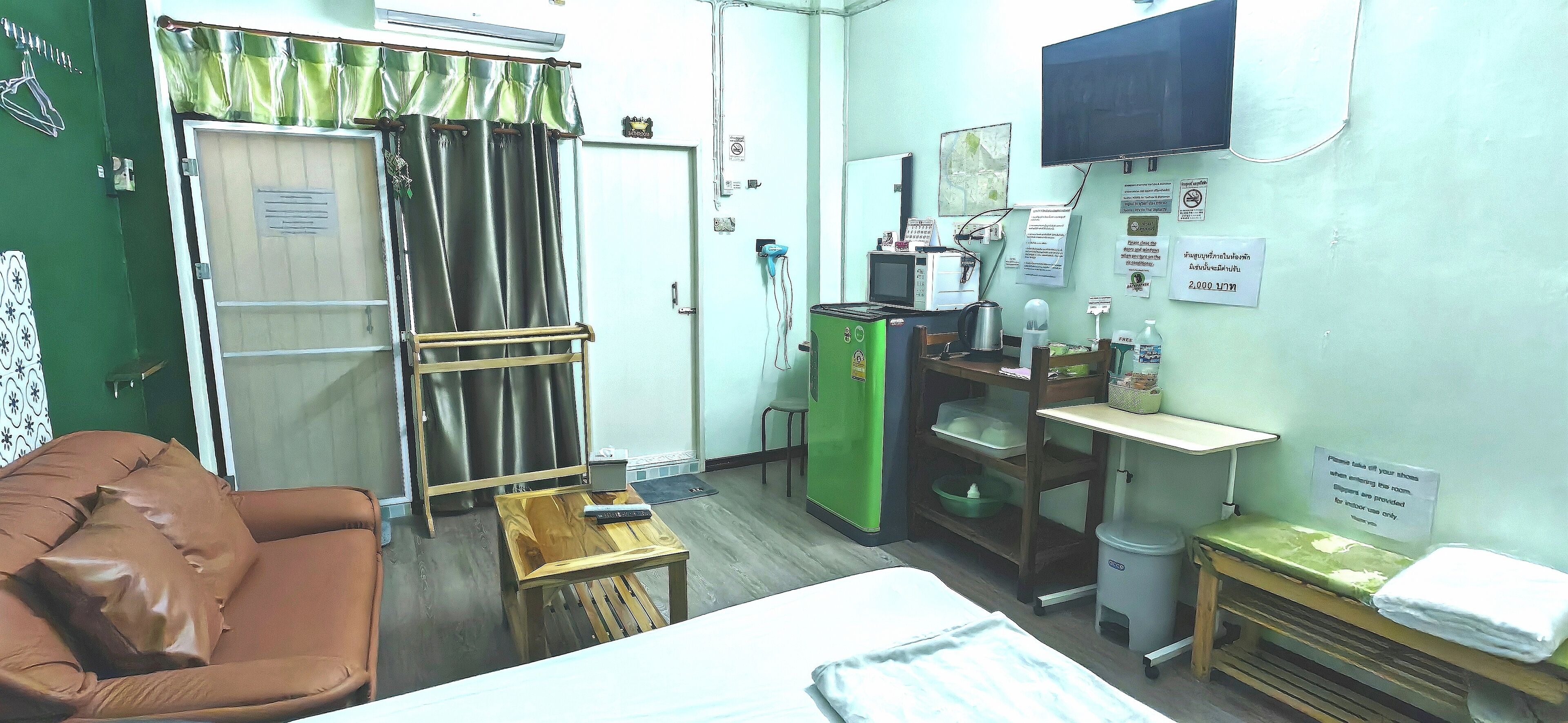 Une petite pièce, de taille modeste, comprenant un lit, une table en bois, une armoire verte et un téléviseur fixé au mur.