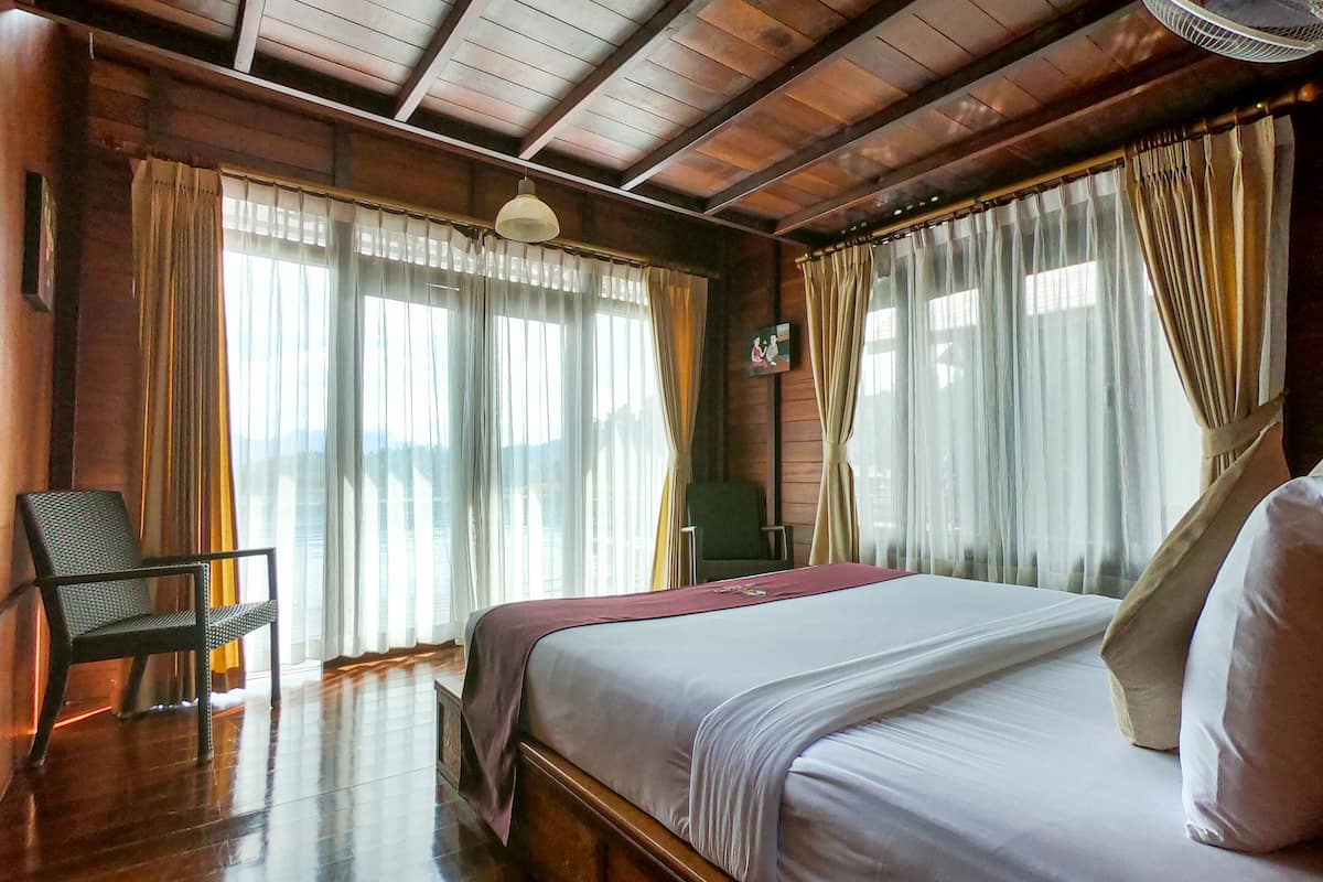 Ein Zimmer mit Holzbalkendecke, einem Bett mit weißer Bettwäsche, einem Stuhl und einem Ventilator.
