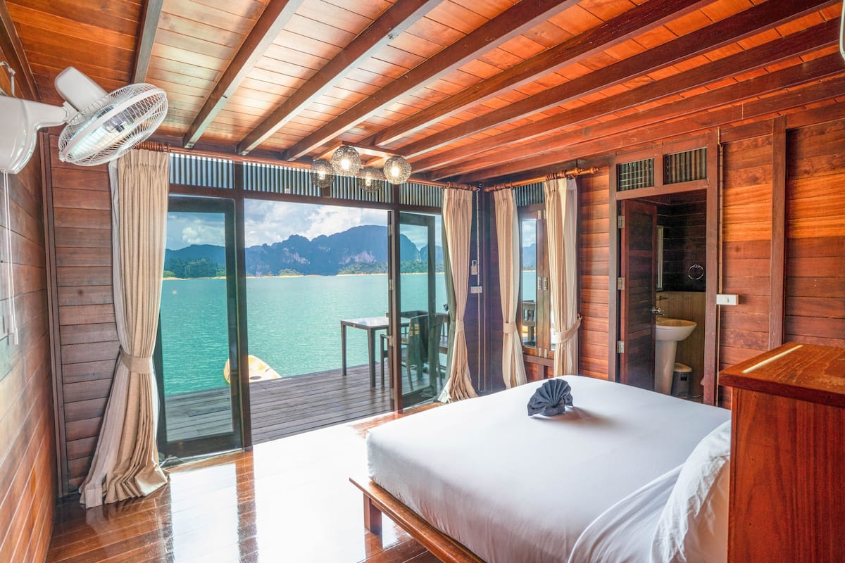 Ein Zimmer in einer Holzhütte mit Bett, Deckenventilator und Meerblick, sowie ein Badezimmer mit Waschbecken.