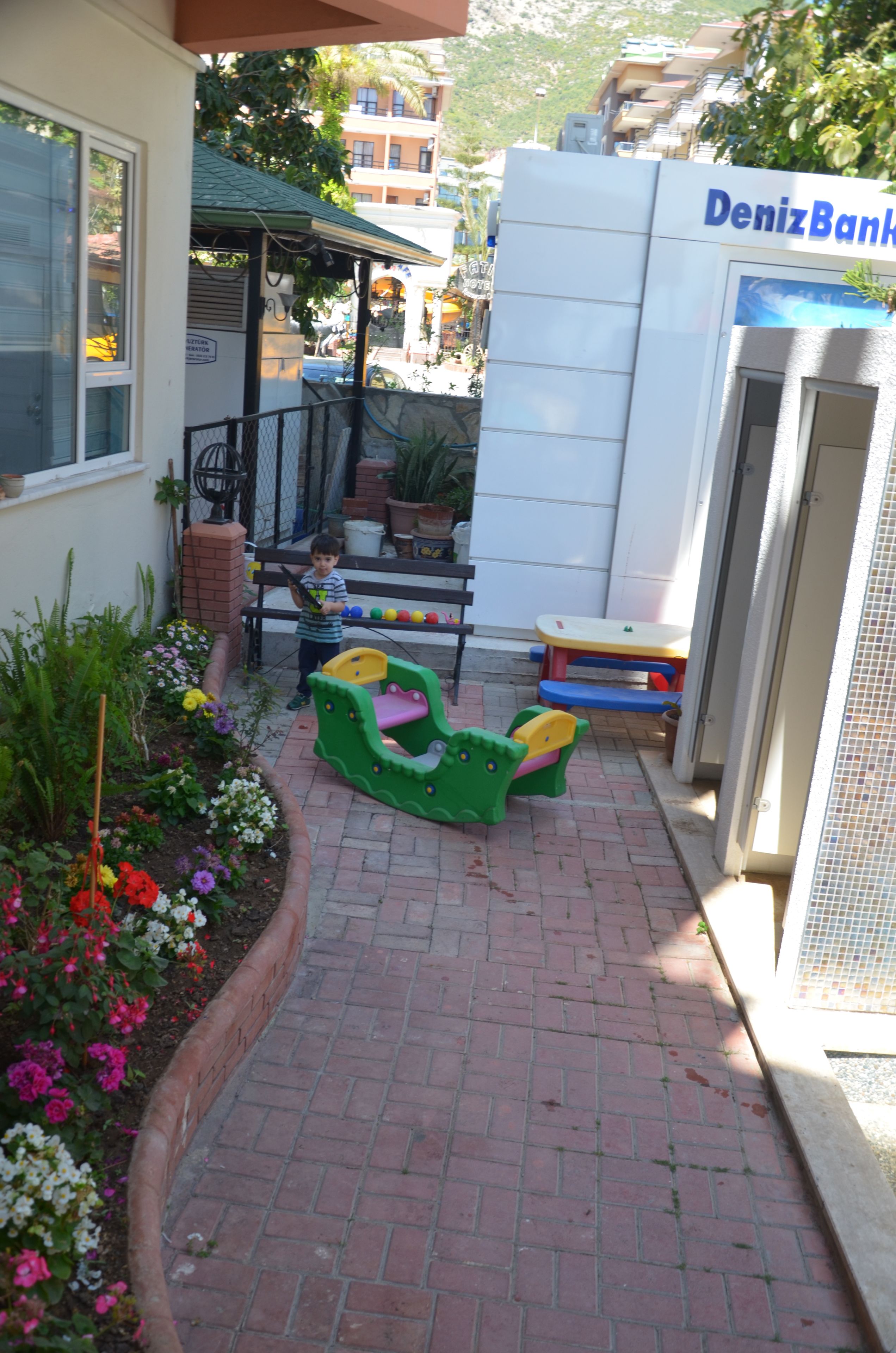 A paved pathway with a bench and children's play area, a building with a sign reading "DenizBank," and a flower bed with various plants.