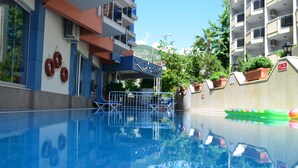 Una piscina con superficie d'acqua cristallina e di colore blu, circondata da un edificio con balconi e un cartello con su scritto "HOTEL".
