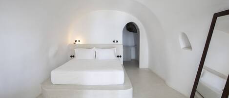 Een minimalistische slaapkamer met een bed centraal onder een gewelfde plafond, een kamerbreed tapijt en een deur naar een andere kamer.