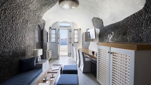 A modern interior with a stone wall, a wooden dining table, a sofa, and a kitchen area with a counter and cabinets. - Ducato di Oia - Adults Only (Santorini)