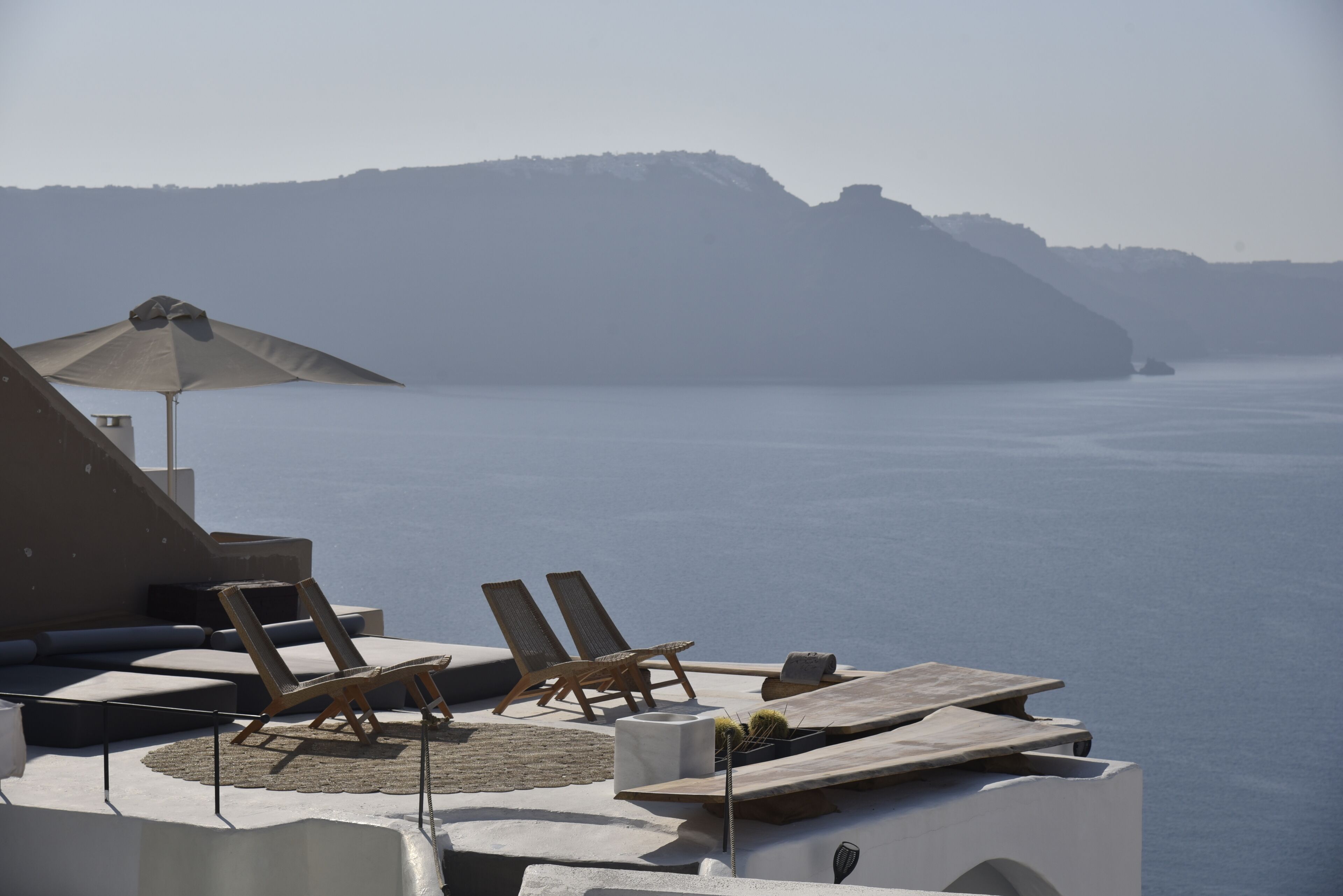 Une terrasse en bord de mer, dotée de chaises longues et d’une table, offrant une vue sur une mer calme et des collines lointaines.