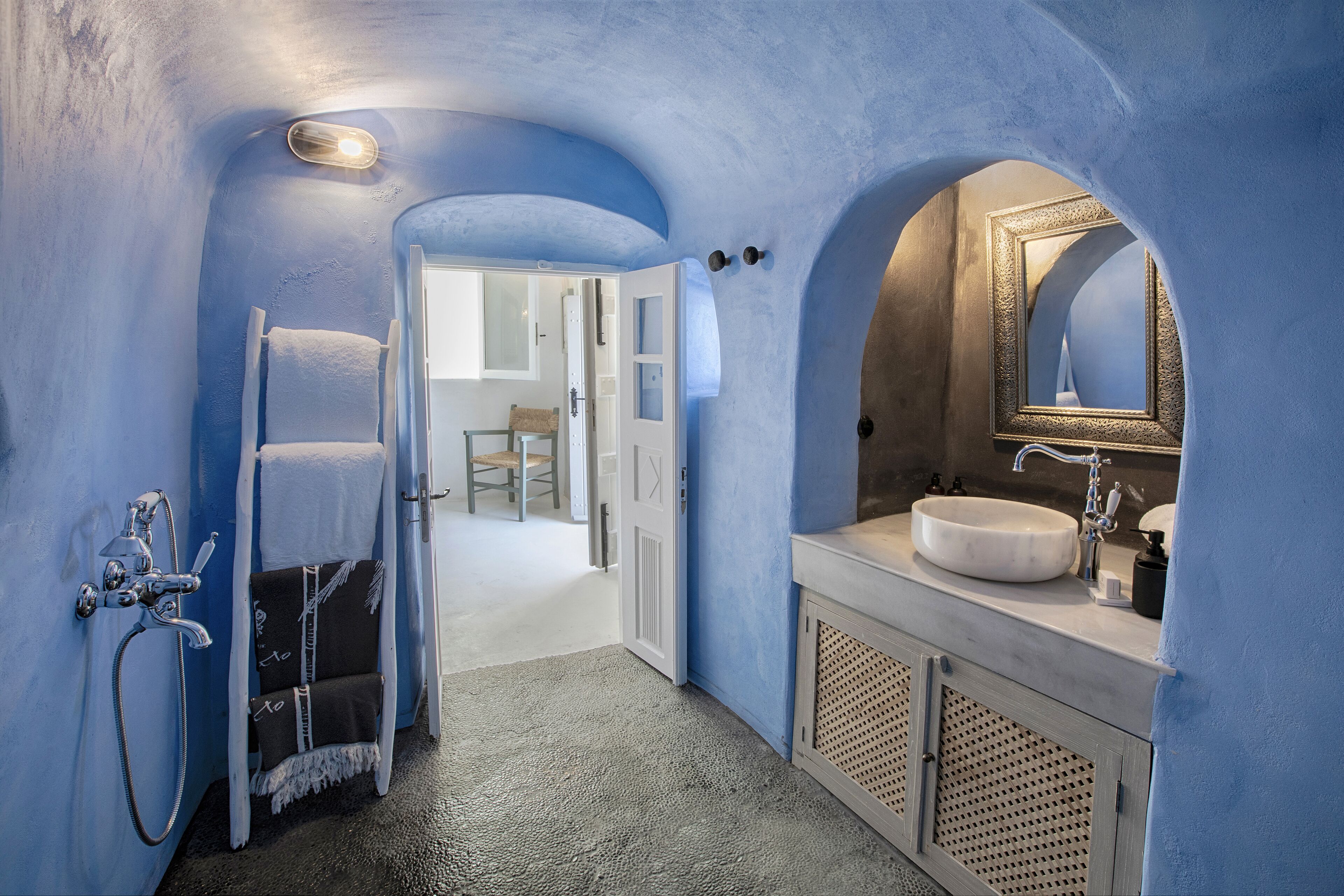 Un pasillo estrecho con pared azul, una puerta blanca que da a una habitación y un baño con lavabo y espejo.