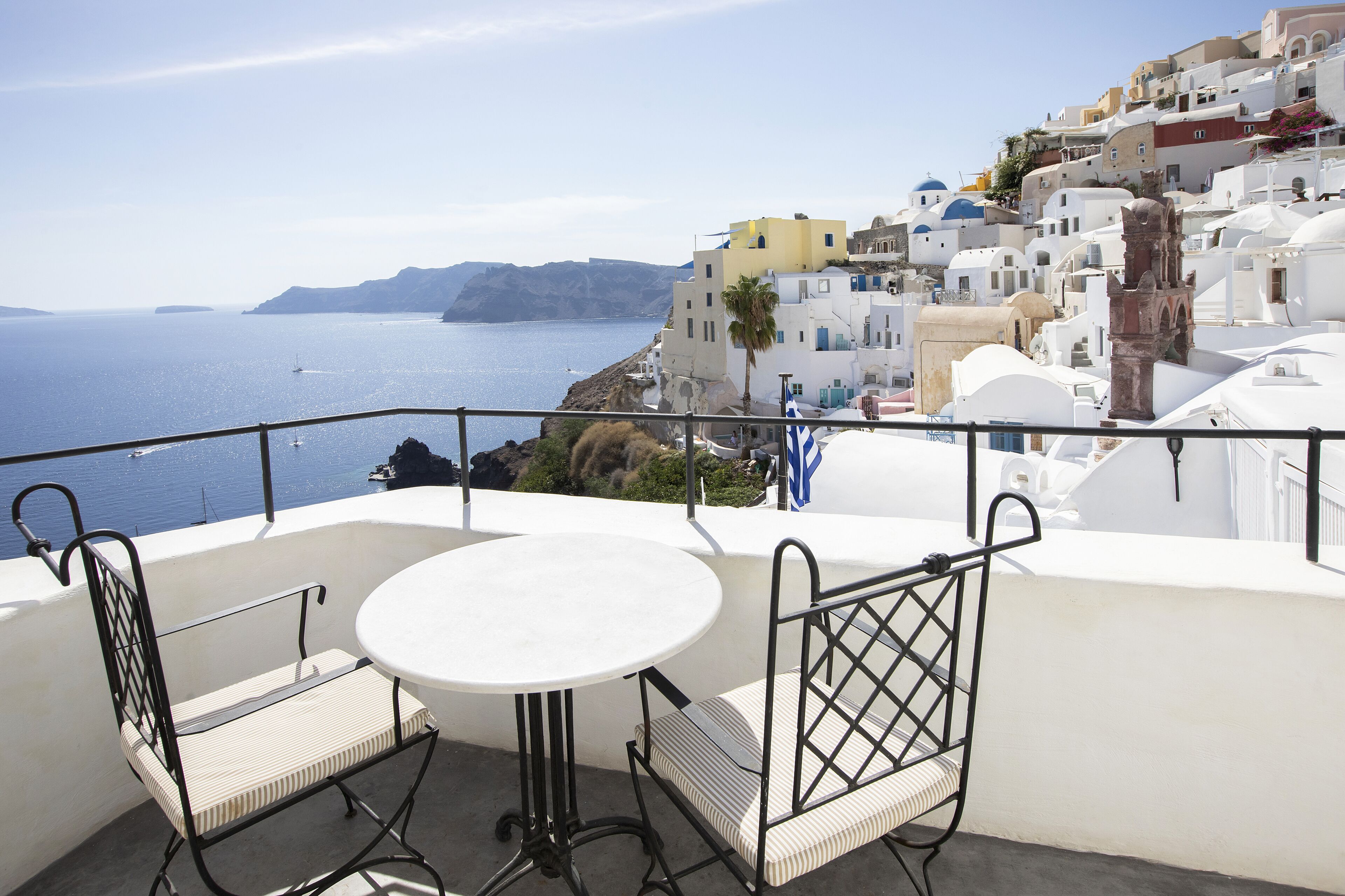 A balcony with a round table and two chairs, overlooking a coastal town with white buildings and a clear view of the sea.