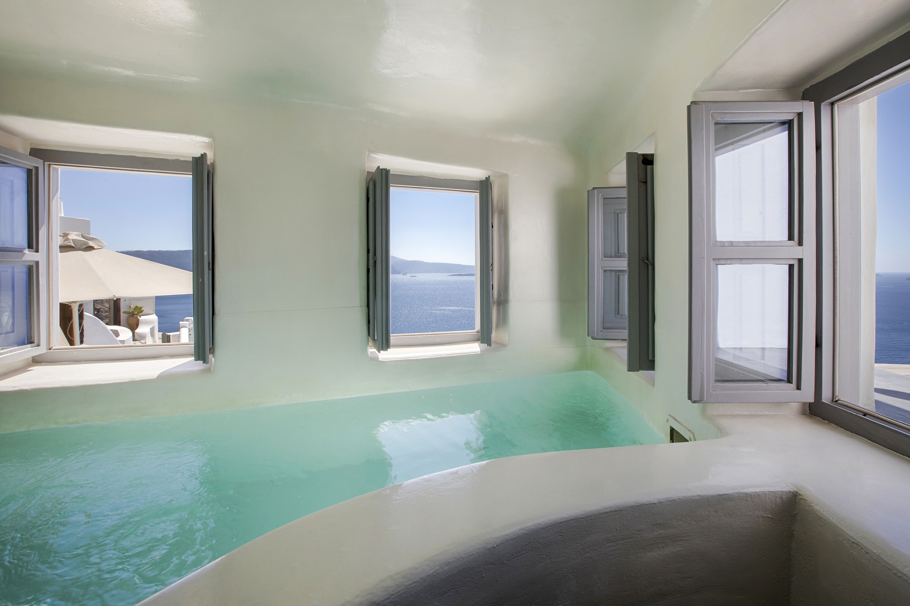A room with a large bathtub, two windows with ocean views, and a small balcony with a table and chairs.