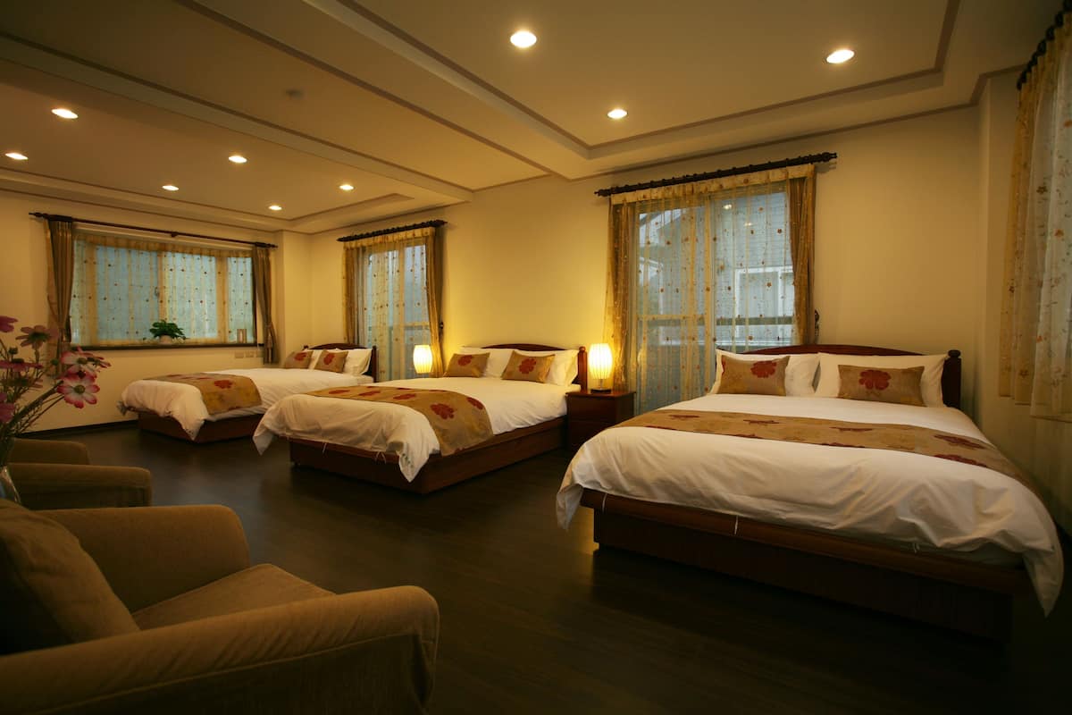 A hotel room with three beds, a sitting area with a chair, and large windows with curtains.