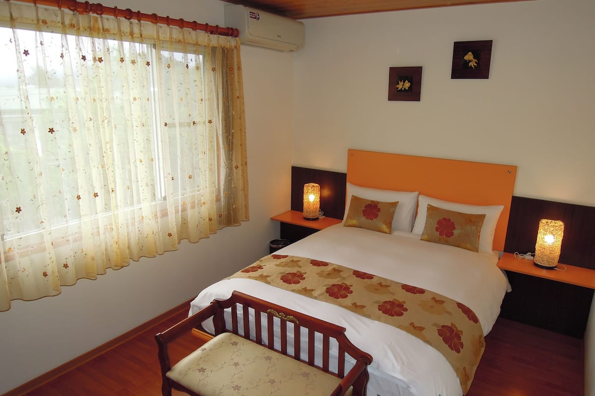 A bedroom with a wooden bed, floral bedding, a wooden chair, two bedside tables with lamps, and a window with floral curtains.