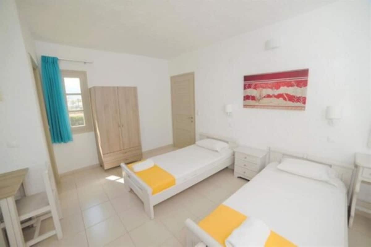 A bedroom with two single beds, a wooden wardrobe, a window with a blue curtain, and a wall-mounted light fixture.